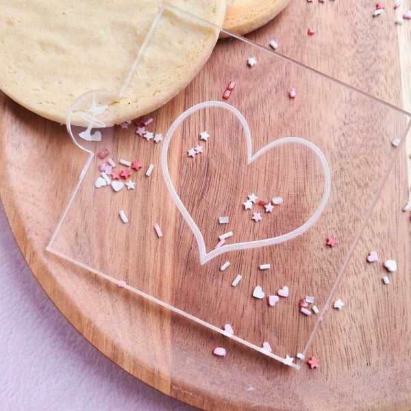 Icing stamp with heart design shown on a wooden plate with sprinkles and cookies