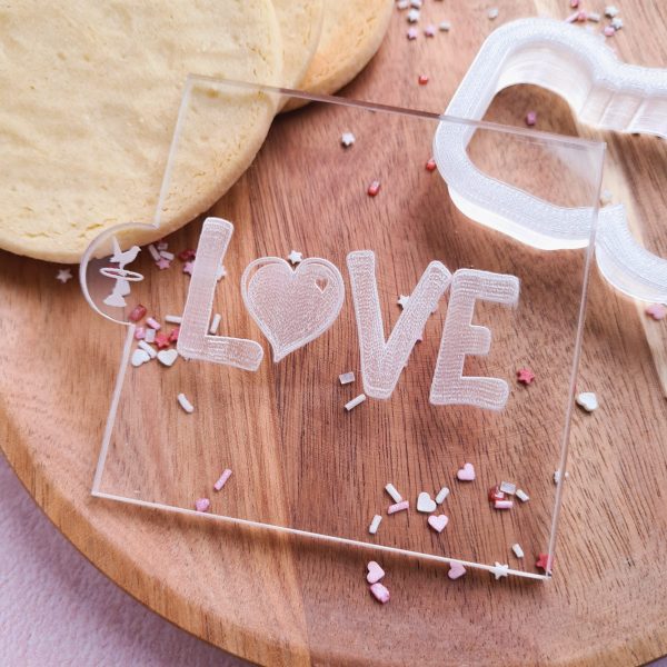 Embosser with LOVE design with a heart for the O, shown on a wooden plate with cookies, sprinkles and a cookie cutter